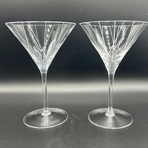 May include: Two clear martini glasses with etched linear designs. The glasses have a triangular bowl shape, slender stems, and round bases. The etched lines radiate from the center of the bowl towards the rim, creating a decorative pattern.