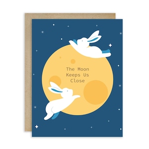 May include: Greeting card featuring a whimsical illustration. Two white rabbits with blue accents appear to be floating near a large yellow moon against a dark blue starry background. The text on the moon reads "The Moon Keeps Us Close."