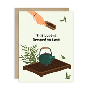 May include: Greeting card with a light green background and a brown border. The card features a teal teapot on a wooden tray, a hand holding a scoop of tea leaves, and the text "This Love is Brewed to Last."