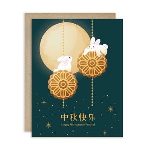 May include: Greeting card with a teal background featuring a full moon and two white rabbits perched on mooncakes. The card includes the text "Happy Mid-Autumn Festival" in English and Chinese characters, with gold star accents.