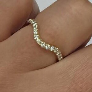 Op de afbeelding: Een goudkleurige ring met een golvend, gebogen bandontwerp. Het oppervlak van de band is bezet met een doorlopende rij kleine, heldere, sprankelende steentjes die de golvende vorm van het metaal volgen, wat zorgt voor een verfijnde uitstraling.