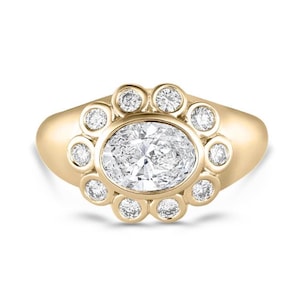 Könnte beinhalten: Ein Goldring mit einem großen, oval geschliffenen, klaren Edelstein in der Mitte, umgeben von einem Kranz aus zehn kleineren, rund geschliffenen, klaren Edelsteinen. Die Steine sind in eine polierte Goldschiene mit glatter Oberfläche gefasst.