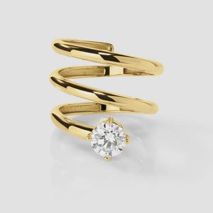 May include: A gold spiral ring featuring a polished metallic band that wraps around in a coil design. The end of the band is set with a single, round, clear, faceted gemstone that sits prominently at the front of the piece, catching the light.