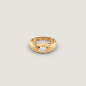 Può includere: Un anello a fascia in oro lucido con una singola piccola pietra preziosa trasparente a forma di cuore, incastonata a filo al centro della spessa superficie metallica arrotondata. L'anello presenta una finitura liscia e riflettente.