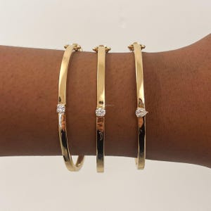 Puede incluir: Tres brazaletes dorados con un diseño sencillo. Cada brazalete presenta una pequeña gema transparente. Los brazaletes se muestran en un brazo, mostrando su estilo elegante y minimalista.