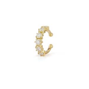 May include: A gold ear cuff featuring a series of clear, round gemstones. The cuff curves around the ear, with a small ball at each end. The gemstones are evenly spaced along the front of the cuff.