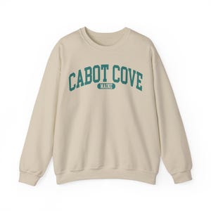 May include: A beige sweatshirt with a teal blue arch design that reads "CABOT COVE MAINE".