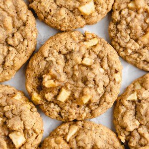 May include: Close-up of several apple oatmeal cookies. The cookies are golden brown with visible pieces of apple and oats. They are arranged on a light-colored surface, showcasing their texture and ingredients.