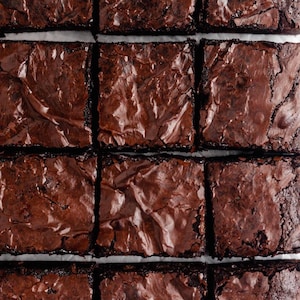 May include: Overhead shot of a grid of freshly baked brownies. Each square brownie has a shiny, crinkled top and a rich, dark brown color. The brownies are cut into neat squares, with white parchment paper visible between them.