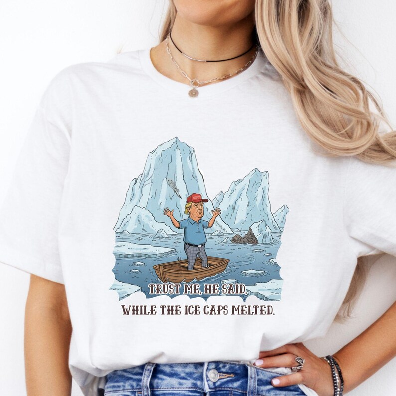 May include: White t-shirt featuring a cartoon illustration of a person in a boat surrounded by melting icebergs. The person is wearing a red cap and blue shirt. Text reads: "Trust me, he said, while the ice caps melted."
