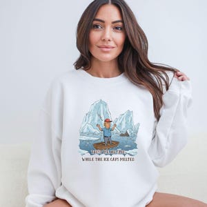 May include: White sweatshirt featuring a cartoon illustration of a man in a boat amidst melting icebergs. The image includes the text "TRUST ME HE SAID" and "WHILE THE ICE CAPS MELTED."