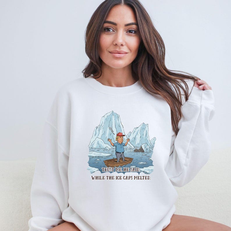 May include: White sweatshirt featuring a cartoon illustration of a man in a boat amidst melting icebergs. The image includes the text "TRUST ME HE SAID" and "WHILE THE ICE CAPS MELTED."