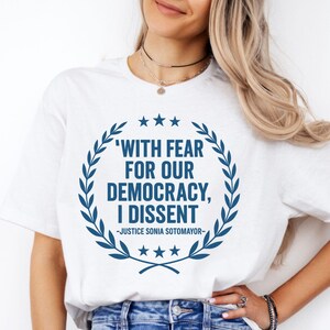 Può includere: T-shirt bianca con un design circolare blu con la citazione "WITH FEAR FOR OUR DEMOCRACY, I DISSENT" attribuita al giudice Sonia Sotomayor. Il design include stelle e una corona d'alloro.