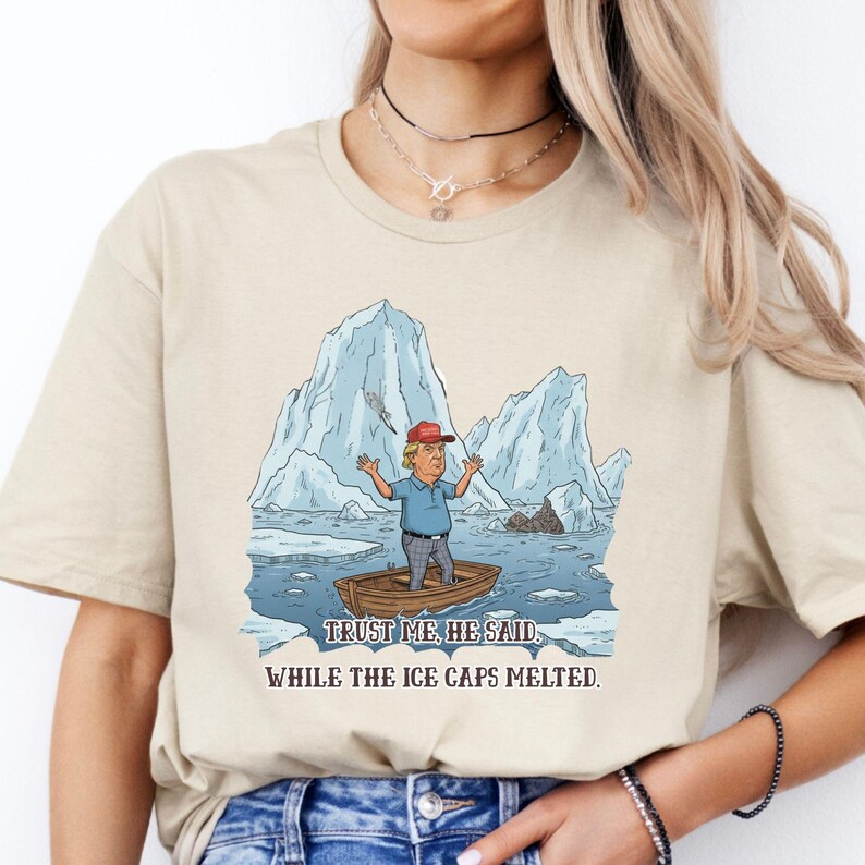 May include: Beige t-shirt with a cartoon illustration of a man in a boat surrounded by melting icebergs. The man is wearing a red cap and blue shirt. Text reads: "Trust me, he said, while the ice caps melted."