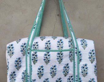 May include: White quilted tote bag with a blue floral pattern. The bag features green and white striped handles and trim. The bag is rectangular with a top zipper closure.