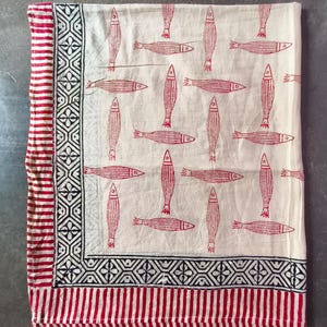 May include: A rectangular textile featuring a cream-colored base with a repeating pattern of red fish. The fabric is bordered by a black geometric diamond motif and an outer edge of red and white vertical stripes, creating a distinct frame.
