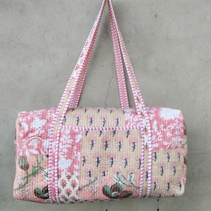 May include: A quilted duffel bag with a floral pattern in shades of pink, white, and green. The bag has two long handles with a striped design. The bag is rectangular with a pocket on the front.
