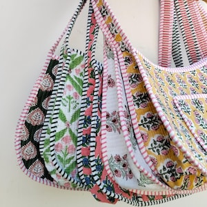 Op de afbeelding: Een collectie gewatteerde hobo-tassen met diverse florale blokprintpatronen in geel, groen, roze en zwart. Elke tas heeft een eigen kleurenschema en is afgewerkt met een roze en wit gestreepte stoffen bies langs de randen en de schouderbanden.