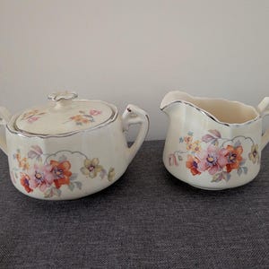 May include: A cream-colored ceramic sugar bowl with a matching lid and a creamer pitcher. Both pieces feature a delicate floral pattern in shades of orange, pink, and yellow, accented with silver trim along the scalloped edges and handles.
