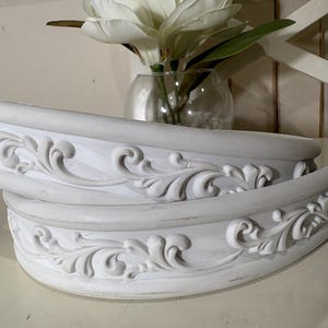 以下が含まれることがあります： 白く湾曲した2つの装飾的な棚板で、精巧な浮き彫りの渦巻き模様が施されています。白い花と緑の葉が入った透明なガラスの花瓶が棚板の後ろにあります。背景は明るくニュートラルな色です。