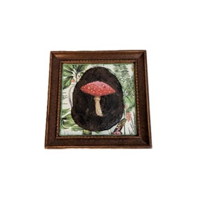 May include: A framed artwork featuring a red and white spotted mushroom against a dark background. The mushroom is centered, with a light brown stem. The background has a botanical print with green leaves and flowers. The frame is a dark brown color.