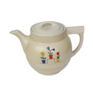 May include: Cream-colored ceramic teapot with a lid and handle. The teapot features a floral design with yellow, blue, and red flowers in pots. The teapot has a spout for pouring and is designed for brewing tea.