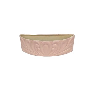 May include: A light pink, half-moon shaped ceramic planter. The planter features a detailed, raised floral design on the front. The interior is a light beige color. The planter is designed to be mounted on a wall.
