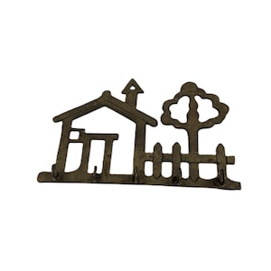 May include: Bronze-colored key holder shaped like a house with a tree and picket fence. The house has a chimney and door. Three hooks are below the house and fence for hanging keys. A decorative item for home organization.