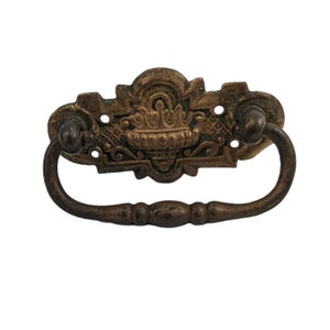 May include: Antique brass drawer pull with an ornate design and a loop handle. The handle is a dark brown color and the design is a lighter brass color.