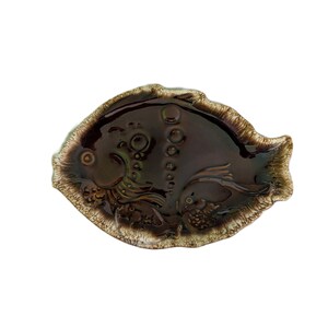 May include: A fish-shaped ceramic dish with a glossy dark brown glaze. The surface features embossed details of two fish and bubbles, with a textured, light-colored rim that contrasts with the smooth, dark center of the decorative tray.