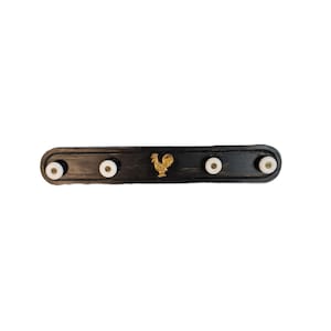 May include: A black wooden coat rack with five white pegs and a gold rooster emblem. The rack is oval-shaped with rounded ends. The rooster is centered on the rack.