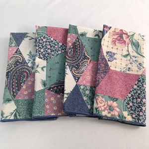 May include: Four folded cloth napkins with a patchwork design. Each napkin features a mix of floral, paisley, and geometric patterns in shades of pink, blue, green, and cream. The edges are finished with a blue trim.