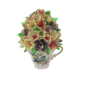 May include: A decorative teacup filled with a bouquet of artificial flowers. The flowers are in shades of cream, pink, and purple, with green leaves. The teacup has a gold handle and a painted bird and floral design.