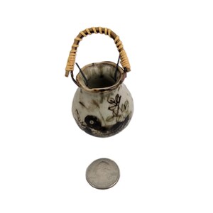 May include: Small, hand-painted ceramic vase with a woven handle. The vase is off-white with brown and green floral designs. The handle is made of woven, light brown material. A coin is next to the vase for size comparison.