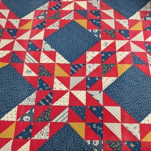 May include: A patchwork quilt with a repeating pattern of red, white, blue, and yellow triangles and squares. The quilt features a central diamond shape in a dark blue floral print, surrounded by red triangles and smaller squares.