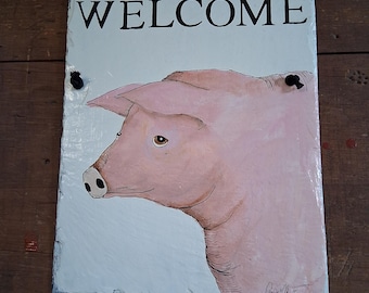 May include: A rectangular slate sign with the word "WELCOME" in black capital letters. Below the text is a pink pig illustration. The sign is hanging by a black cord. The background is a wooden surface.