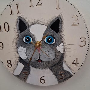 May include: A round wall clock featuring a hand-painted cat design. The cat has gray and white fur, blue eyes, and a pink nose. The clock face is cream-colored with black numbers and a dotted border. The clock hands are gold.