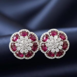 May include: A pair of flower-shaped earrings. Each earring features a central cluster of small white stones surrounded by a ring of oval-cut, deep red stones. The earrings are set against a dark blue background.