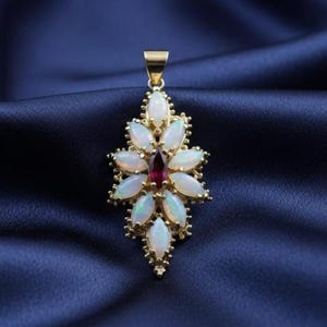 May include: A gold pendant featuring a floral design with white opal petals surrounding a central red gemstone. The pendant has a gold bail and is displayed against a dark blue background.
