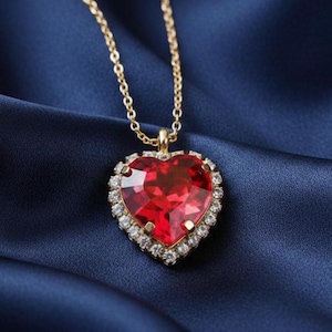 May include: A gold-colored necklace with a heart-shaped pendant. The pendant features a large, faceted red gemstone surrounded by a halo of small, clear crystals. The necklace is displayed on a dark blue, silky fabric.