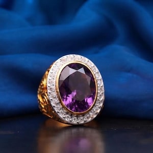 Pode incluir: Um anel de ouro com uma grande ametista roxa de corte oval no centro. A pedra é rodeada por um halo de pequenas pedras brancas transparentes cravadas numa armação prateada. A banda de ouro apresenta padrões intrincados em relevo.