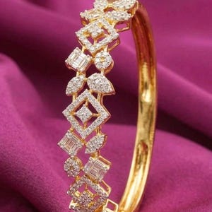May include: A gold bangle bracelet with a diamond design. The bracelet features geometric shapes, including squares, rectangles, and circles, all encrusted with small diamonds. The bracelet is set against a deep purple background.