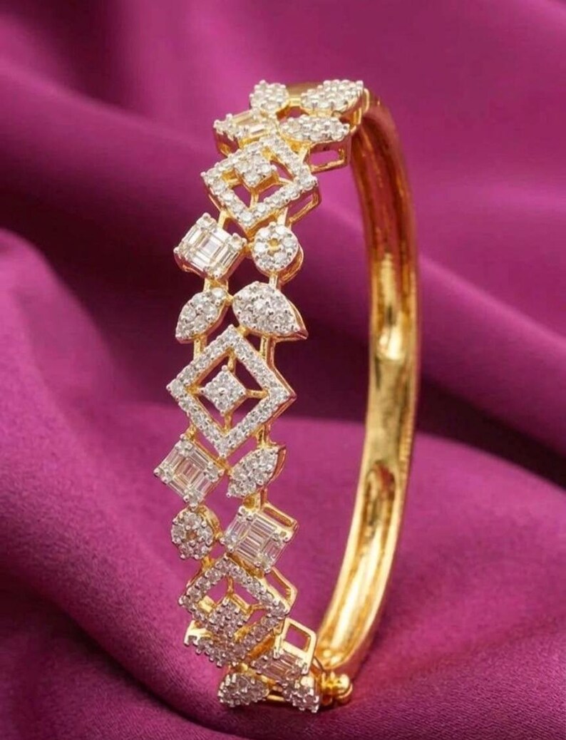 May include: A gold bangle bracelet with a diamond design. The bracelet features geometric shapes, including squares, rectangles, and circles, all encrusted with small diamonds. The bracelet is set against a deep purple background.