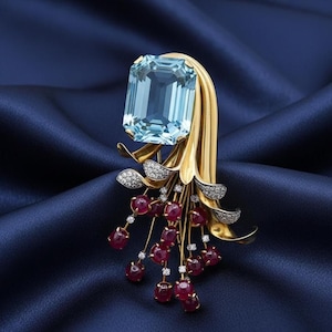 May include: A gold brooch with a large, rectangular, light blue gemstone. The brooch has a floral design with ruby-coloured accents and small diamond details. The background is a deep blue fabric.
