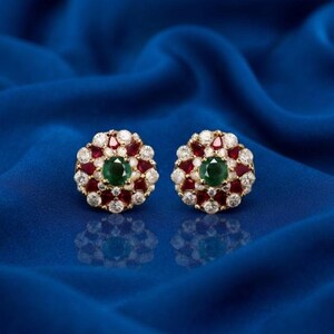 May include: A pair of gold-tone stud earrings featuring a central round-cut green gemstone surrounded by a halo of alternating pear-cut red gemstones and round-cut clear white gemstones arranged in a floral-inspired cluster pattern.