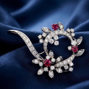 May include: A silver brooch with a floral design, featuring clear stones and red gemstones. The brooch has a curved shape with a decorative pin. The jewellery is set against a deep blue background. The brooch is a decorative accessory.