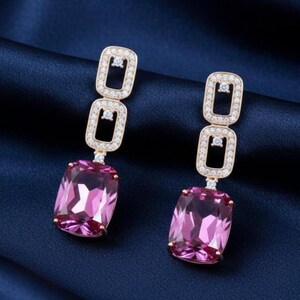Può includere: Un paio di orecchini color oro con un pendente rettangolare in pietra preziosa rosa. Gli orecchini presentano due forme rettangolari impilate incastonate con piccoli cristalli trasparenti sopra la pietra preziosa rosa.