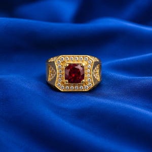 May include: A gold ring featuring a central square-cut deep red gemstone surrounded by a halo of small clear stones. The band has a wide, rectangular face with additional clear stones set into the shoulders in a triangular pattern.