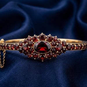 May include: A gold bracelet featuring a central oval-cut, deep red gemstone, encircled by smaller red gems. The hinged bracelet is set against a dark blue backdrop.