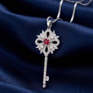 May include: A silver-toned necklace featuring a key-shaped pendant. The pendant head is adorned with marquise and round clear stones surrounding a central round red stone. The key shaft is also encrusted with small clear stones on a chain.